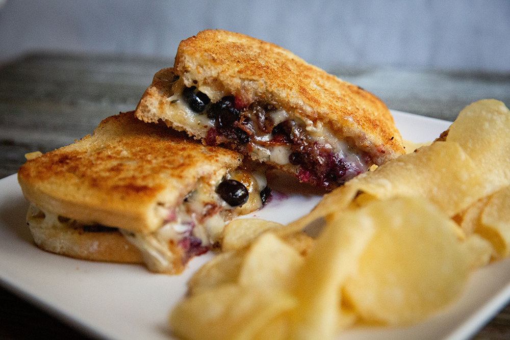 Grilled Sandwich filled with Blueberries a soft brie cheese and bacon with a side of crispy kettle chips on a white plate. 
